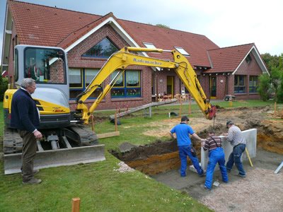 Gelber Minibagger bei Erdarbeiten im Garten eines Wohnhauses, daneben drei Arbeiter