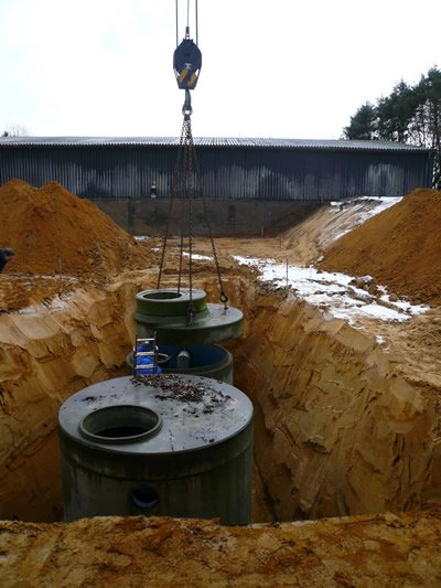 Betonbehälter hängt am Kranhaken in einer tiefen Sandgrube vor einer Halle, davor ein bereits gesetzter Behälter
