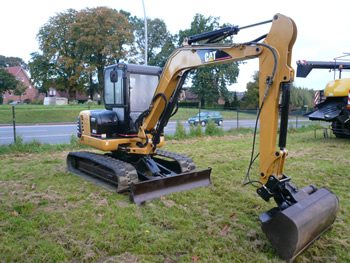 Gelber CAT-Minibagger auf Gummiketten mit Planierschild auf einer Wiese an einer Straße, dahinter Bäume und Häuser