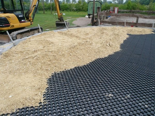 Minibagger verteilt hellen Sand über schwarzen Gitterelementen, die auf der Kreisfläche ausgelegt sind