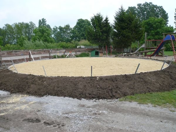 Fertig aufgebauter kreisrunder Roundpan mit heller Sandtretschicht und umlaufendem Erdwall, im Hintergrund ein Spielplatz mit Rutsche