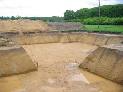 Große ausgehobene Baugrube mit abgeböschten Sandwänden, Pfützen auf der Sohle und einer Leiter an der hinteren Wand
