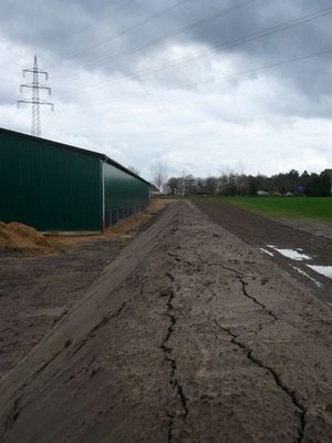 Frisch planierte Erdfläche mit Trockenrissen entlang einer grünen Halle, darüber Strommast und bewölkter Himmel