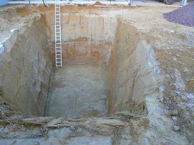 Tiefe, senkrecht abgestochene Baugrube in hellem Sand mit einer Aluminiumleiter an der Wand
