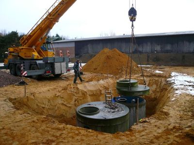 Autokran hebt einen runden Betonbehälter in eine Baugrube, in der bereits ein zweiter Behälter steht