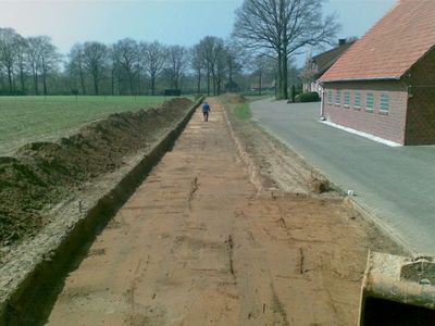 Langer, flacher Aushubstreifen neben einer Landstraße und einem Backsteinhaus, am Ende geht eine Person