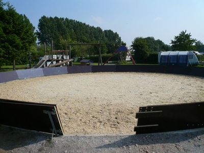 Runder Reitplatz mit Sandboden und dunkler Bretterbande, im Hintergrund Bäume und Spielgeräte
