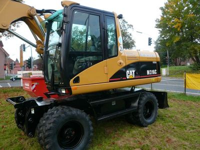 Gelber CAT-Mobilbagger M316C auf Gummireifen auf einer Grünfläche an einer Kreuzung