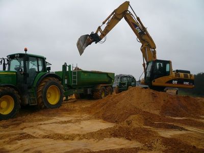 CAT-Kettenbagger 320C belädt auf einer Sandfläche einen grünen Muldenkipper hinter einem Traktor