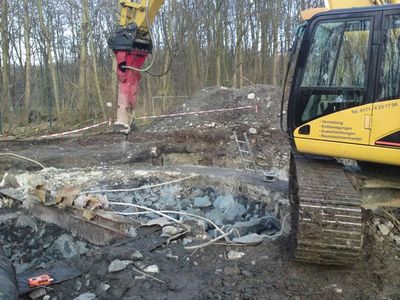 Kettenbagger mit Hydraulikhammer zerkleinert Betonreste, abgesperrt mit Flatterband vor einem Wald
