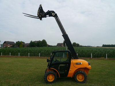 Kompakter Teleskoplader mit ausgefahrenem Arm und Palettengabel auf einer Wiese vor einem Maisfeld
