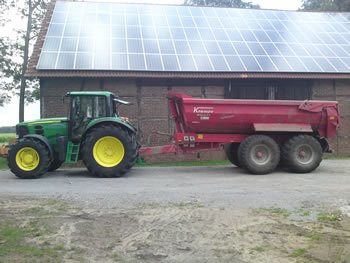 Grüner Traktor mit rotem Halfpipe-Kipperanhänger vor einer Scheune mit Solardach