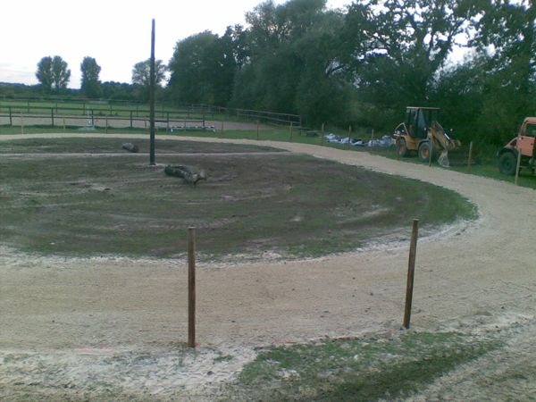 Ovalbahn im Bau: eine Rundbahn führt um eine begrünte Innenfläche, ringsum stehen Holzpfosten, im Hintergrund Baumaschinen