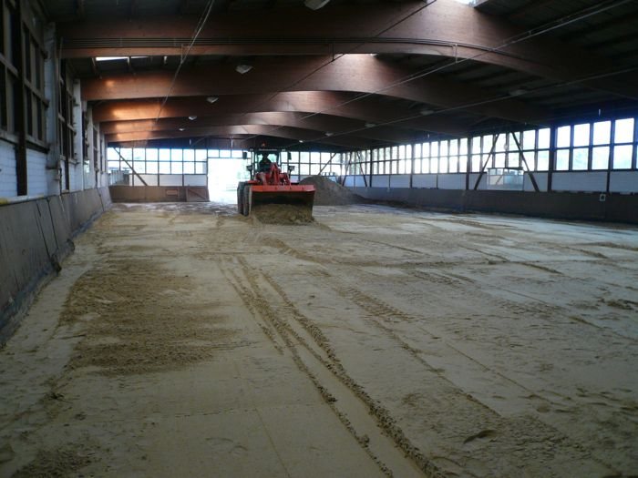Blick in eine Reithalle mit Holzdachstuhl, im Hintergrund schiebt ein oranger Radlader Sand über die Fläche