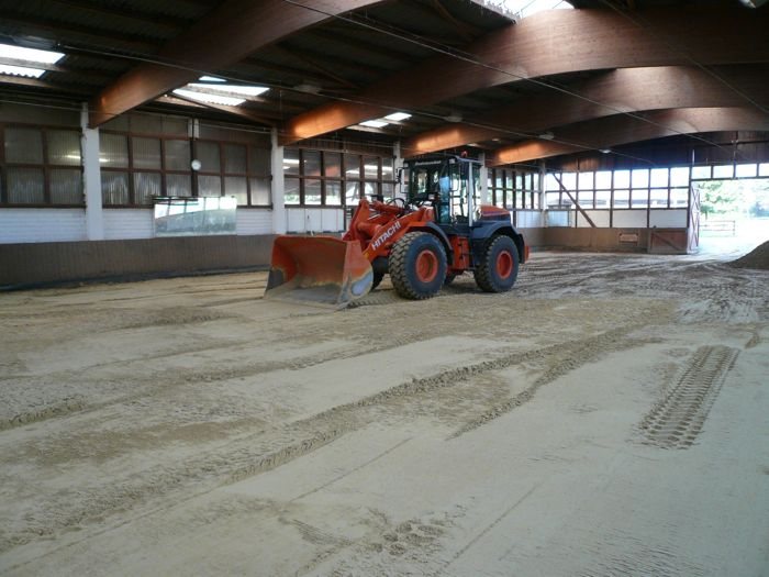 Oranger Radlader mit abgesenkter Schaufel auf dem Sandboden einer Reithalle