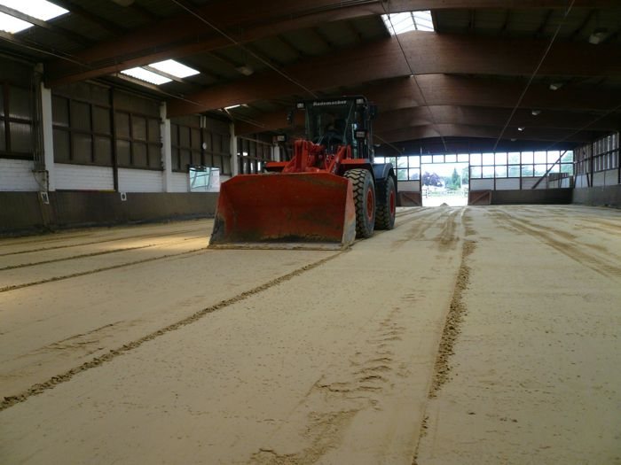 Radlader schiebt Sand in einer Reithalle, im Boden sind Reifenspuren zu sehen