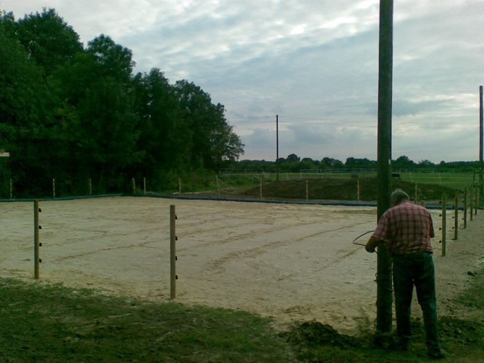 Abgezogener Sandplatz mit Pfosten und Drahtzaun, im Vordergrund ein Mann an einem hohen Holzmast, dahinter Weiden